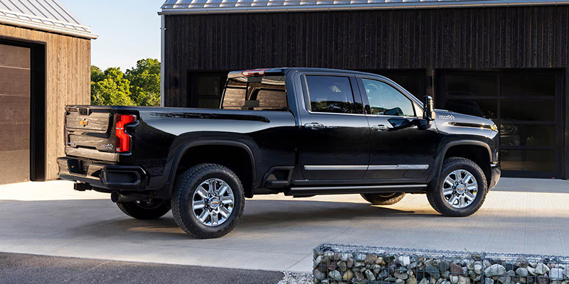 A black pickup truck is parked on a concrete driveway in front of a dark wooden building. The scene is bright and clear, conveying a modern and rugged aesthetic.