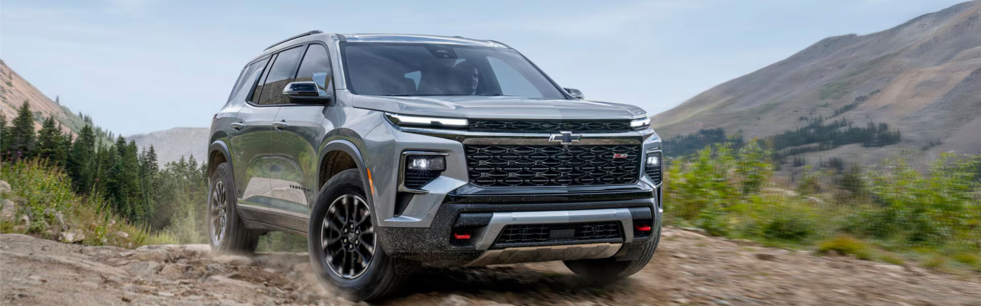 2026 Chevrolet Traverse SUV driving off-road on a rocky path, surrounded by trees and mountains.