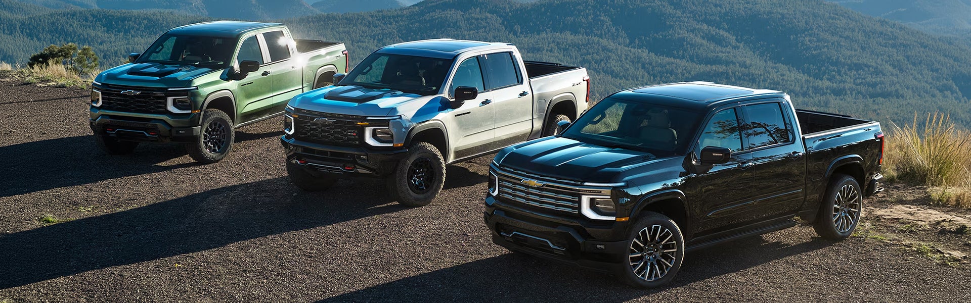 Three Chevrolet Silverado 1500 trucks in different colors parked on a mountain, showcasing their rugged design against a scenic backdrop.
